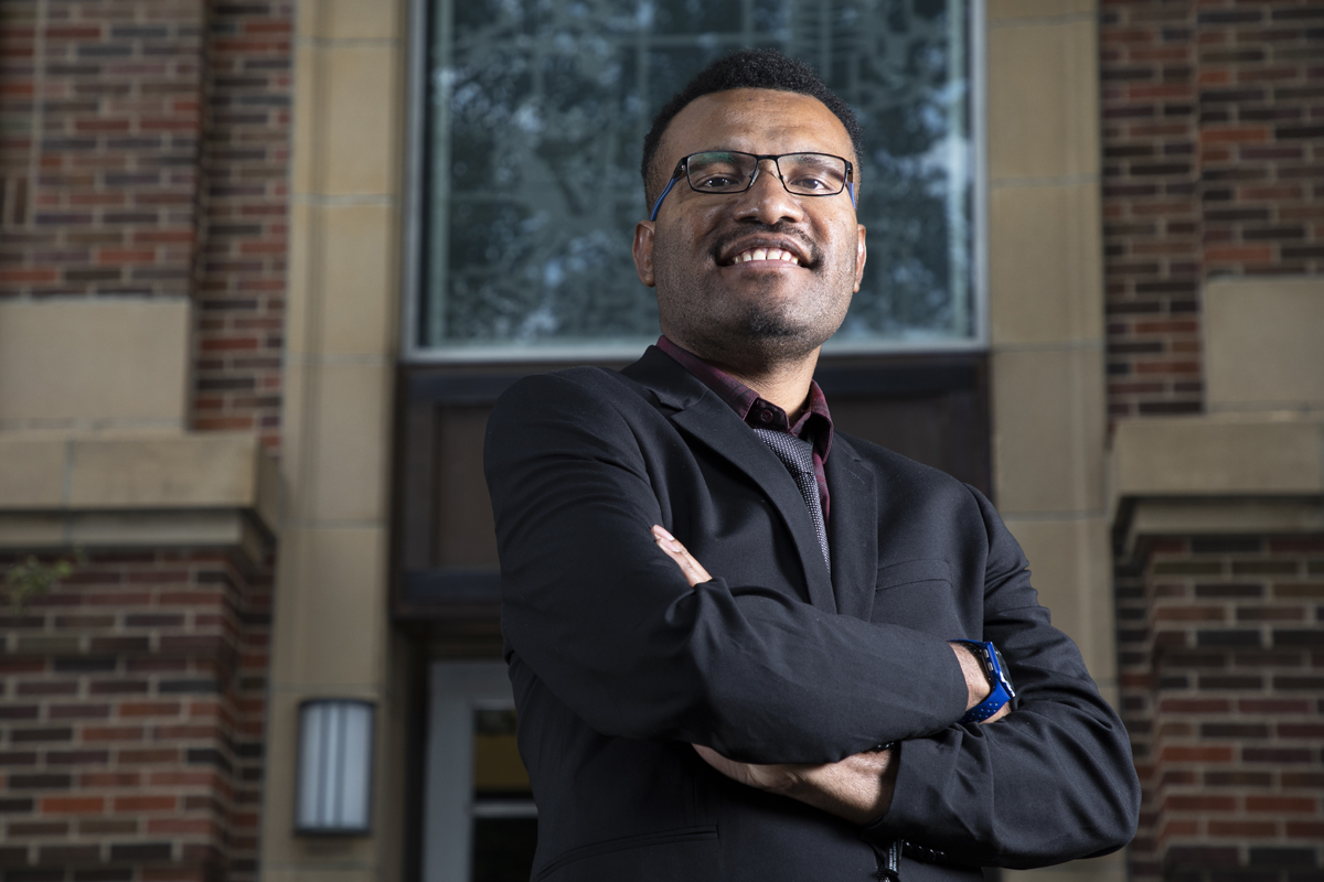 Yulianus Kogoya stands in front of a building at UM with his arms folded over his chest and smiles at the camera